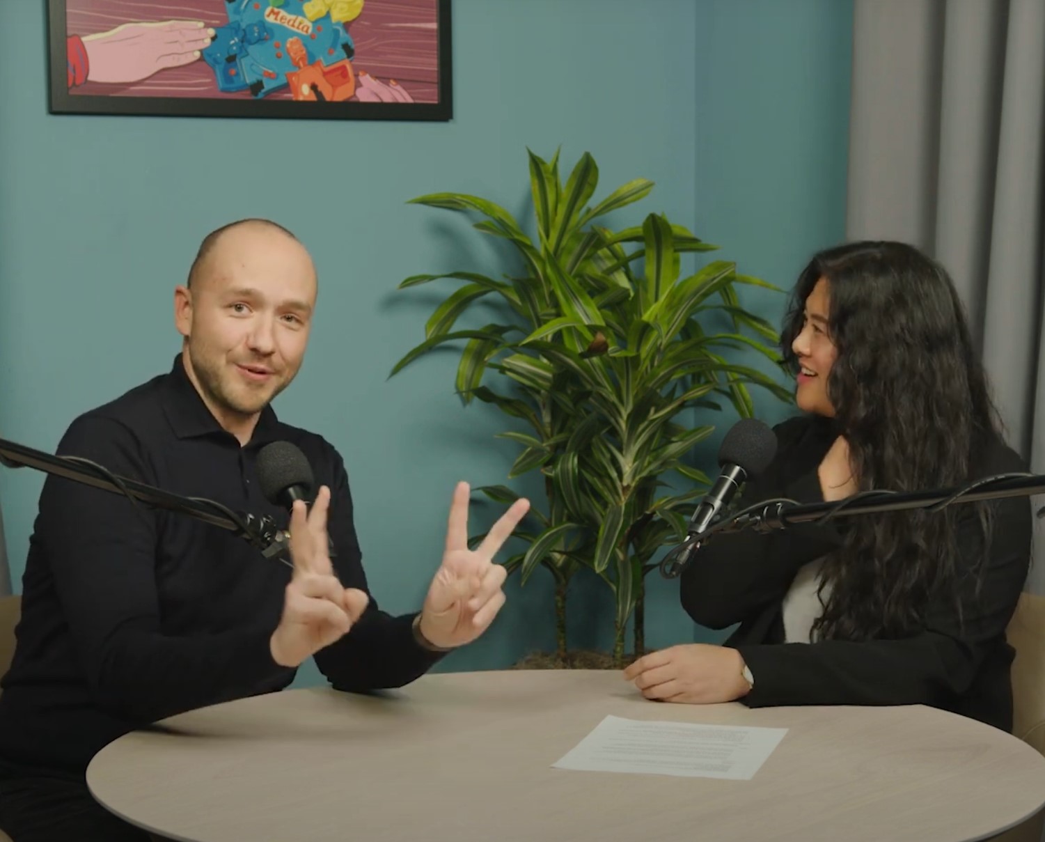 A man and a woman sit at a table in front of microphones. The man is smirking and making quotation marks with his fingers.